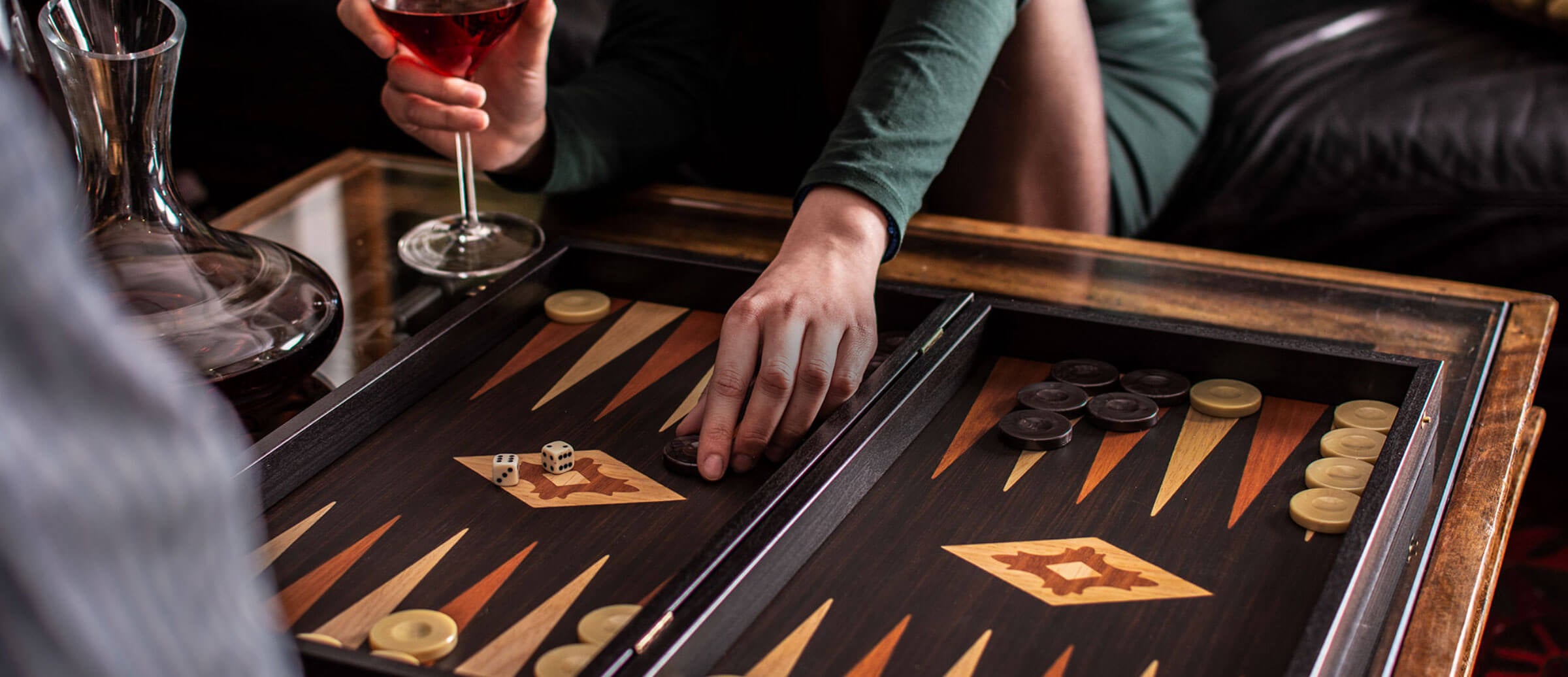 Person playing backgammon while enjoying a glass of red wine in a cozy, elegant setting with a wooden game board.1621243260e1af0c20-0